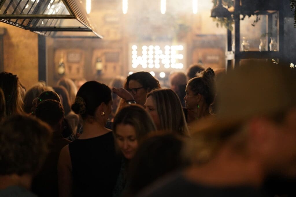 A crowd of people at a event, in the distance a lit sign with the word wine